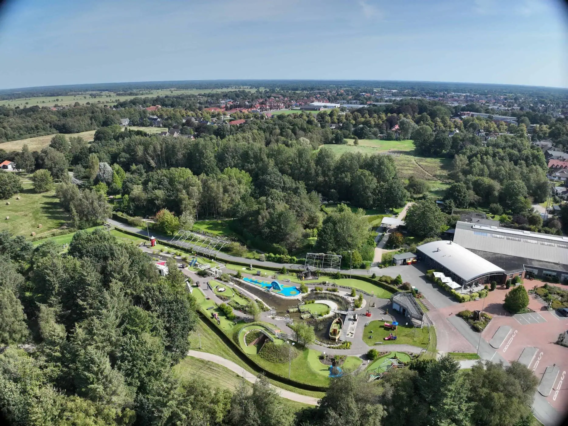 Luftaufnahme eines Parks mit Spielplatz, Spazierwegen und umgebendem Grün. Im Hintergrund sind Gebäude und eine entfernte Stadtlandschaft zu sehen.