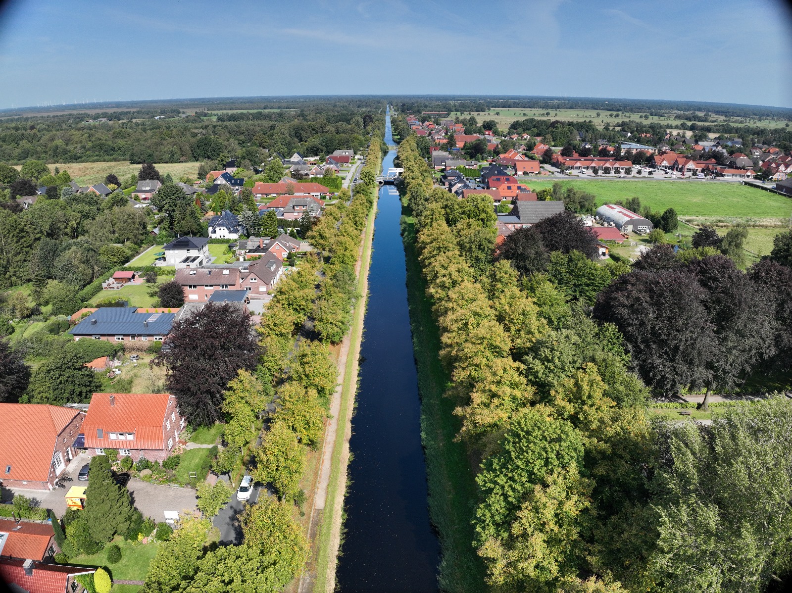 Luftaufnahme eines schmalen, von Baumreihen umgebenen Kanals, der eine kleine Stadt mit Häusern auf beiden Seiten teilt, eingebettet in eine ländliche Landschaft unter einem klaren blauen Himmel.